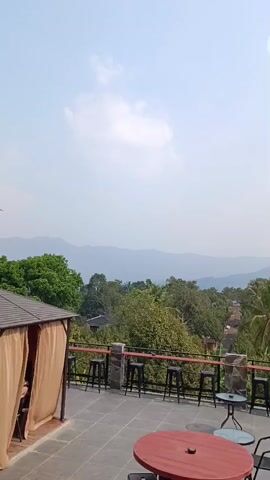 The video shows a panoramic view from a high vantage point, likely a rooftop or balcony, overlooking a lush, green landscape with mountains in the distance. The foreground features an outdoor seating area with tables and chairs, suggesting a cafe or restaurant. A red round table is in the center, with several black bar stools arranged along a railing. A covered seating area with tan curtains is visible on the left. The sky is bright and appears to be a slightly hazy day. The overall impression is one of a serene and scenic outdoor dining or relaxation spot.