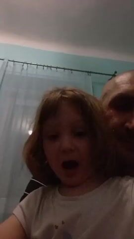 A young girl with brown hair is held in someone's arms. Her head and upper torso are visible in the foreground. She is wearing a white t-shirt and is facing the camera and talking. A bald man’s face is partially visible over her shoulder in the background. The background appears to be a room with a teal wall and a sheer white curtain hanging behind them. The lighting is somewhat dim.