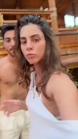 A woman in a white shirt films herself and a shirtless man, who is wearing white shorts. They are dancing outside and the camera spins during the dancing. There is sand on the ground around them, and a building made of wooden planks is in the background. There are also sandals and bottles on the ground nearby.