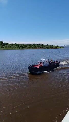 A boat travels through a body of water on a sunny day. The boat is gray and white with a red deck. Trees line the distant shore, and the sky is blue with a few clouds. The water is a dark color, reflecting sunlight. As the boat moves, it leaves a wake behind it. It passes a red and white navigational marker in the water. The camera angle is from above and to the side of the boat.