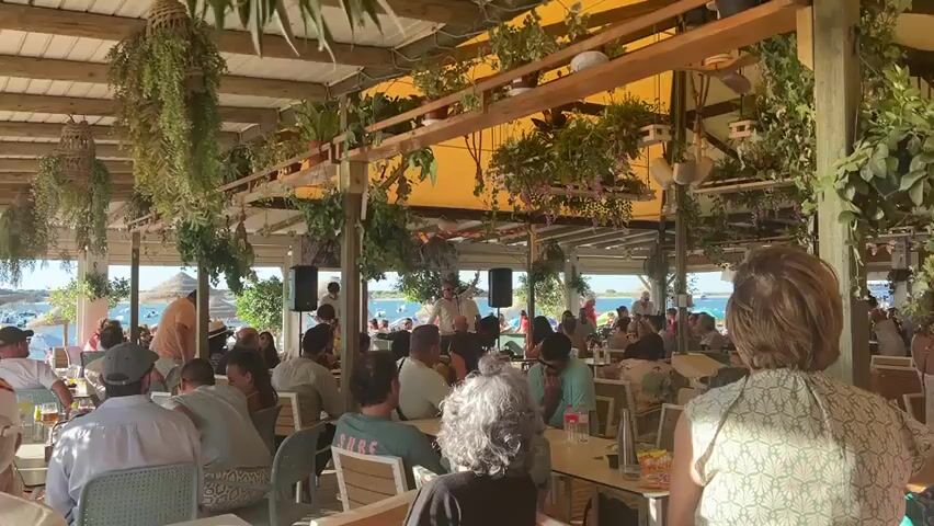The video shows a wide shot of an outdoor restaurant with a thatched roof, decorated with hanging plants. Many people are seated at tables, some eating and drinking, while others watch a performance on a small stage in the distance. A man is standing on the stage holding a guitar, singing and playing. Behind him, there is a body of water, possibly the ocean, with boats and a beach visible. The overall atmosphere appears to be relaxed and festive.