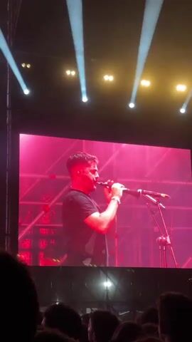 A large screen at a music concert shows a man playing a wind instrument into a microphone. He wears a black shirt and dark pants. The background is red. The stage lighting is bright. The crowd is visible below the screen. The video then shows the man rapping into the microphone as the red background changes.