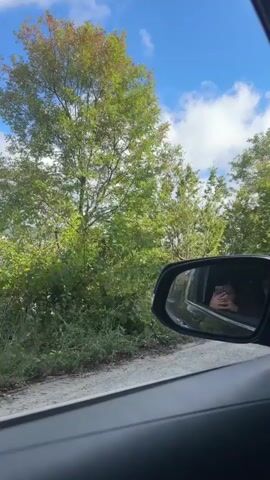 The video begins with a view from inside a vehicle, looking out the passenger window. Lush green trees and foliage line a dirt road, under a bright blue sky with fluffy white clouds. The side mirror of the vehicle is visible, reflecting the face of a person holding a phone, presumably taking the video.

As the video progresses, the camera pans slightly, revealing the interior of the vehicle. The driver's side of the car is visible, including the dashboard and a part of the steering wheel. Through the driver's side window, another vehicle, a white minivan, passes by. Some of the passengers inside the minivan are visible through its windows. The camera continues to move, showing more of the interior, including the headliner and a grab handle. The view outside remains consistent with trees and vegetation.