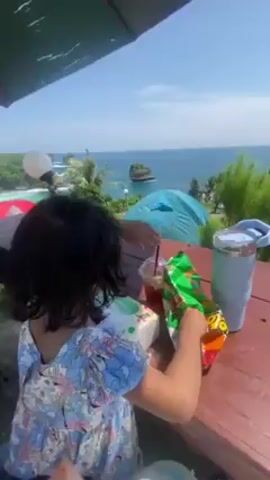 A wide-angle shot shows a young girl sitting at a red picnic table, opening a bag of chips on a bright day. She is wearing a blue and white floral dress. On the table are tissues, a drink with ice, and a light blue tumbler. The camera pans to the right, revealing a view of a grassy cliffside with tents pitched near the edge. The ocean is visible in the background, with a small rocky island in the distance. Several people are standing near the tents, and there are bushes and small trees in the foreground. The scene is framed by a green umbrella overhead. The sky is blue with a few clouds.