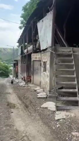 Okay, here's a detailed description of the video:

**Setting:** The video appears to be shot in a rural or semi-rural area. The ground is dirt and gravel, with signs of recent rain or moisture.

**Structures:** There is a weathered building constructed from stone and wood. The building has a two-story section. The upper level is open, seemingly a porch or balcony with wooden supports. The roof is a mix of corrugated metal and wood. An exterior stone staircase leads up to the second story on the right-hand side of the shot.

**Objects:**
*   A metal door is visible on the side of the building.
*   Stone pavers are used to create a pathway leading to the door.
*   A metal fence or corrugated metal sheeting is attached to part of the building.
*   A utility pole and wires are visible in the background.
*  What appears to be a small light is on the pole.

**Animals:**
*   A flock of chickens is gathered near the building and under the overhanging structure.

**Overall Impression:** The scene has a rustic, possibly impoverished feel. The building appears to be aged and possibly in need of repair. The overall environment suggests a rural or agricultural setting.