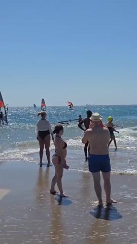 The video shows a crowded beach with several people windsurfing in the ocean under a clear blue sky. The windsurfers are scattered across the water, some closer to shore and others further out. On the beach, several individuals are visible, some walking along the shoreline and others standing near the water's edge. The water appears choppy, indicating windy conditions suitable for windsurfing. The overall atmosphere suggests a recreational day at the beach with various activities taking place.