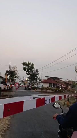 The video captures a scene at a railway crossing. A barrier, painted with alternating red and white stripes, is lowered, indicating that a train is expected or passing. On the right side of the frame, a person is seated on a motorcycle, facing away from the camera. The person's hand is on the handlebar. In the background, across the railway tracks, there are buildings, trees, and power lines. Several other motorcycles and possibly some cars are stopped at the crossing, also waiting. The sky is overcast and appears to be either early morning or late afternoon. A sign with the text "AS SAID" is visible on a pole to the right of the frame. The overall impression is a temporary pause in traffic due to the railway crossing.