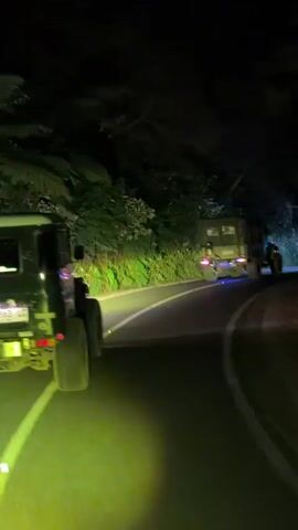 The media file depicts a nighttime convoy of off-road vehicles driving on a winding, unlit road through a forested area. In the foreground, a black Mercedes-Benz G-Class, possibly modified, is seen from the rear. It has bright yellow headlights that illuminate the road ahead and cast a green glow on its tires and the road surface. The license plate is visible and reads "R 1863 LC".

Following behind the lead G-Class are two other similar vehicles, also appearing to be off-road SUVs, with their headlights on. The second vehicle is a darker color, and the third vehicle is a lighter blue. The road is paved and has white lane markings. The surrounding environment is dark, with trees and foliage lining the sides of the road, suggesting a rural or mountainous location. The time of day is clearly night due to the darkness and the use of headlights.

The activity captured is the vehicles driving in a procession, likely on an adventure or journey. The overall mood is adventurous and possibly a bit mysterious due to the nighttime setting and the isolated road. There are no people clearly visible within the vehicles. The location context provided is Tosari, Indonesia, which aligns with a forested, mountainous terrain often found in that region.