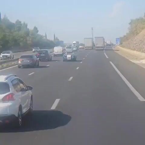 The video shows a traffic jam on a highway. A blue and white convertible car with white stripes is in the middle lane. A person is driving the convertible. Other vehicles, including trucks and vans, are also visible in the traffic jam. The weather appears to be sunny and clear. The video is shot from another vehicle.