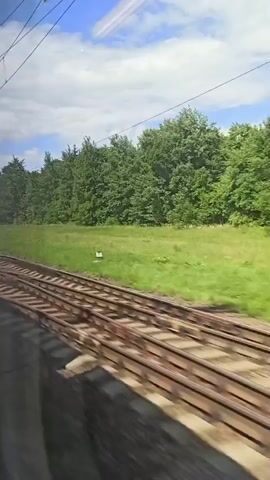 The video shows a view from a moving train, looking out the window. The train tracks are visible in the foreground, running parallel to the direction of travel. To the left of the tracks, there is a grassy field with scattered trees. In the distance, a dense forest can be seen. Overhead, there are power lines and a cloudy blue sky. The train appears to be moving through a rural or suburban area, with occasional utility poles and possibly distant buildings visible. The lighting suggests it is daytime.