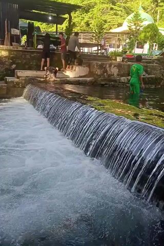 A waterfall flows over a stone wall into a pool of water. In the background, there are people sitting and standing, and trees are visible. The scene appears to be a natural setting with some man-made elements.