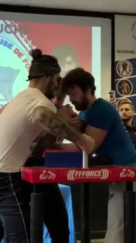 Two men are engaged in a competitive arm wrestling match. The man on the left wears a white t-shirt, black pants, and a bandana, and has prominent tattoos on his left arm. The man on the right wears a blue shirt and grey sweatpants. Their hands are clasped together on a red arm wrestling table with the logo "FFFORCE" displayed on its surface. A judge stands between them, observing the match. Behind them, a projector screen displays a presentation of sorts, while on the right, a poster showcases various disciplines. In the background, two spectators watch, one of whom is recording the match with his phone. The scene appears to be taking place indoors, possibly in a sports club or training facility.