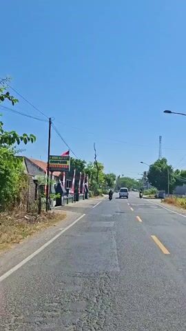 This is a vertical video shot from a moving vehicle on a paved road. The sky is clear and blue. On the left side of the road, there is a building with a red tile roof, a fence, and trees. There are also several Indonesian flags displayed on poles. Further down the road on the left, there appears to be a business with signage. On the right side of the road, there are more trees, some buildings, and a tall communication tower. The road has yellow lane markings. A white van is traveling ahead in the same direction, and two motorcycles are also visible on the road. The vegetation is green and lush, suggesting a warm climate. The road surface shows some signs of wear and tear, with visible patches. The overall impression is of a rural or suburban setting on a sunny day.
