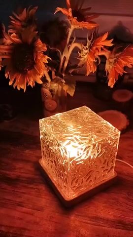 This video shows a square, textured glass lamp that emits a warm, orange glow. The lamp is placed on a wooden surface, likely a table. Behind the lamp, a vase contains several sunflowers with wilted petals. The overall ambiance is cozy and dimly lit. The video is static, with no discernible movement or action.