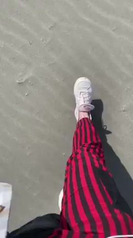 The video shows a person standing on a sandy beach. The sand is light grey and has ripple patterns. The person is wearing white sneakers and black pants with red and black striped fabric on one leg. The video then pans up to show the ocean in the distance under a clear blue sky.  The ocean appears calm with small waves.  The beach appears mostly empty.