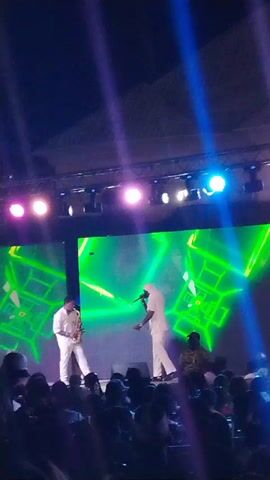 On a stage brightly lit with white, blue, and pink lights, two men dressed in white are performing in front of a crowd. The man on the left plays a saxophone, while the man on the right holds a microphone and appears to be singing or rapping. Behind them is a large screen displaying dynamic green and white geometric patterns. The crowd is visible in the foreground.
