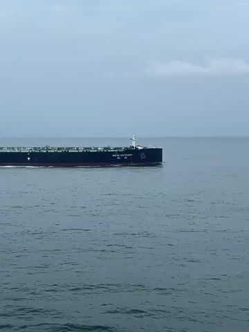 A large tanker ship is sailing on the ocean. The ship is dark colored with white structures on its deck. The name "NEW VICTORY" is visible on the side of the ship. The sea is calm with small waves. The sky is overcast and gray. The camera is moving alongside the ship, showing its full length as it passes.