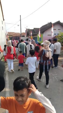 The video captures a group of people, likely participants in an outdoor event, standing in a line. The individuals are of various ages and dressed in casual clothing, with some wearing red tops and white pants, and others in white shirts and black pants. Some are also wearing headscarves, suggesting a cultural context.

The scene appears to be a street or pathway, with houses and buildings visible in the background. Indonesian flags are prominently displayed, indicating a national celebration or event. The ground is paved, with some areas of rougher texture and gravel. The lighting suggests it is daytime, with the sun casting shadows, implying a bright and possibly warm day.

The people are arranged in a line, with some individuals facing forward and others looking towards the side. Their postures and the overall composition of the group suggest they are either waiting for instructions, about to start an activity, or have just finished one. There are no clear indicators of a specific game or competition, but the organized formation hints at a planned event. The presence of a few adults and children together implies a community or family-oriented gathering.

The overall mood appears neutral to positive, with no overt signs of distress or excitement. The focus on the line of people and the surrounding environment creates a sense of community and participation in a local event in Posong, Indonesia. The video, recorded vertically, gives a close-up perspective of the participants and their immediate surroundings.