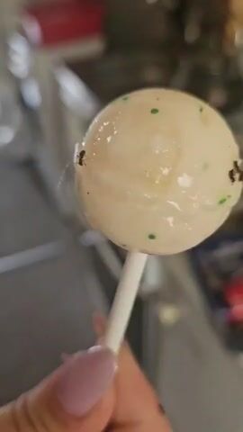 A close-up shot shows a hand holding a white lollipop with green speckles. Several ants are crawling on the surface of the lollipop. The background is blurred, but appears to be an indoor setting, possibly a kitchen.
