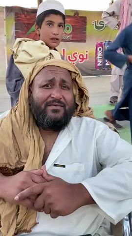 A man in a wheelchair, wearing a white shirt and a tan headscarf, looks off to the side. He has a beard and appears to be speaking. Behind him, a young boy in a white cap and traditional clothing looks towards the same direction as the man. In the background, a banner with the word "NITRO" and images of energy drinks is visible. Several other individuals are standing and sitting, some are wearing traditional attire. The environment appears to be outdoors, possibly at an event or gathering.