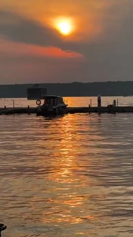 This media file depicts a serene sunset over a body of water, likely a lake or river, in Троицкое, Russia. The sun, a bright orb, is partially obscured by clouds, casting a warm, golden-orange glow across the water. The light reflects off the rippling surface, creating a shimmering path.

In the foreground, a wooden dock extends into the water. A small, dark-colored boat is moored to the dock, its silhouette contrasting with the bright reflection. A life preserver is visible on the boat, suggesting its use for recreational activities. Further back on the water, a larger, dark, rectangular structure, possibly a houseboat or a floating building, is visible. Beyond that, a tree-lined shore forms the horizon.

The overall mood is peaceful and tranquil, indicative of a calm evening. The time of day is clearly sunset, given the intense orange and red hues in the sky and the low angle of the sun. The weather appears fair, with clouds present but not obstructing the sunset's beauty. The presence of the dock and boat suggests a recreational area or a place where people engage in water activities. No visible text is present in the media file.