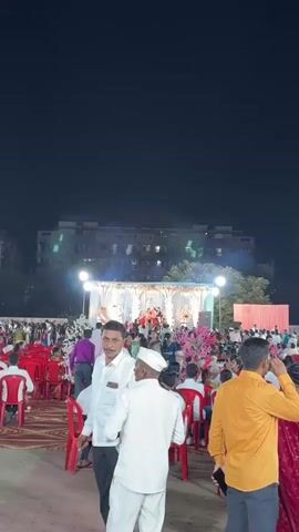 A crowded outdoor wedding celebration is underway. At the center stands a brightly illuminated stage, adorned with elaborate decorations, where a couple is celebrating. The air is filled with the excitement of sparkling fireworks. The crowd, composed of diverse people, is gathered around the stage. Buildings are in the background. The scene captures the festive atmosphere of the event.