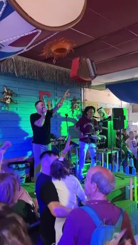 This video captures a lively indoor music event at what appears to be a bar or club in Fuengirola, Spain.

The first shot focuses on a small stage where a live band is performing. On the left, a male singer, wearing a black t-shirt and light-colored pants, stands with a microphone, gesturing to the audience with his right hand. He has tattoos on his arms. To his right, a male drummer, wearing a maroon t-shirt with "OKLAHOMA" written on it and light-colored pants, is actively playing a drum set. In the background, there's a blue wooden wall and various decorative items, including an accordion hanging from the ceiling. Blue stage lights illuminate the performers. Several people are gathered in front of the stage, including one man holding a small child, enjoying the music.

The video then pans across the wider venue, showing a crowd of people dancing and socializing. The ceiling is decorated with numerous large flags, predominantly red, white, and blue, suggesting a festive or Caribbean theme. These flags resemble the flag of the Dominican Republic, among others. The atmosphere is energetic, with many people smiling and moving to the music. Flashing red and blue lights contribute to the party ambiance. In the background, there are signs that appear to read "BONGO BAR," further suggesting the type of establishment.

The overall impression is one of a vibrant evening celebration with live music and dancing.