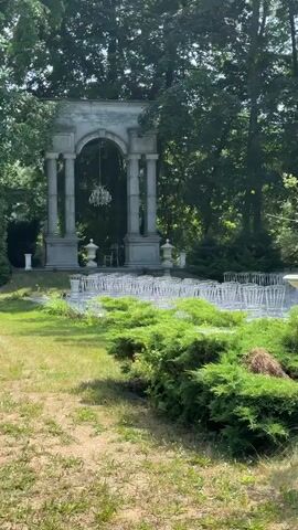 The video shows an outdoor wedding ceremony setup in a garden. A paved walkway lined with white chairs leads towards an ornate stone archway. A chandelier hangs from the archway, and two decorative urns flank the entrance. Lush green trees and bushes surround the area, creating a natural and serene environment. The sun is shining, casting shadows on the ground, indicating it is daytime. There are no people visible in the video. The scene is peaceful and appears to be set for an event.