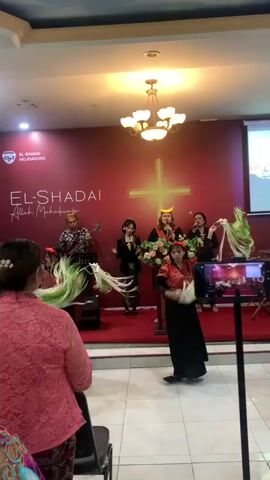 The video opens with a wide shot of a church stage adorned with a large red backdrop featuring the text "EL-SHADAI Allah Mahakuasa." A cross symbol is subtly integrated into the backdrop. On stage, a band plays instruments, with a guitarist and a singer visible. In front of the band, a dancer in traditional attire, including a colorful headdress and flowing green and white sashes, performs. The dancer's movements are energetic, with the sashes creating a dynamic visual effect.

The camera then pans to the right, showing a few audience members seated in the foreground. One woman in a pink lace top is clearly visible from behind. As the video progresses, the camera shifts focus to different parts of the audience and the stage. We see more audience members, some of whom are also dressed in traditional clothing, including a woman with a yellow flower in her hair and another with a gold and red headpiece. The lighting suggests an indoor event, possibly a religious service or cultural performance. The environment appears to be a hall or auditorium, with chairs arranged for seating and plain walls on the sides. The overall atmosphere seems festive and celebratory.