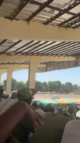The video shows a crowd of people wearing green uniforms and hats under a covered seating area with a metal roof. The crowd is looking towards an open area where a few people are standing near green and yellow tents. One person in the foreground is holding a booklet and appears to be reading or speaking. The camera moves slightly, showing more of the crowd and the open area beyond. The day appears sunny.