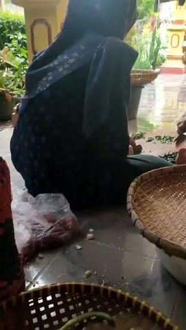 The video shows a person's hands chopping carrots on a cutting board. In the foreground, there is a woven basket filled with green beans, and next to it, another woven basket holds shallots and garlic. To the left, there is a red woven basket. In the background, a person wearing a dark blue hijab and gray pants is visible, sitting on the floor. There is also a plastic bag with some red items inside, and a gas cylinder is partially visible. The scene appears to be an outdoor or semi-outdoor area with tiled flooring and some potted plants. The overall impression is of food preparation.