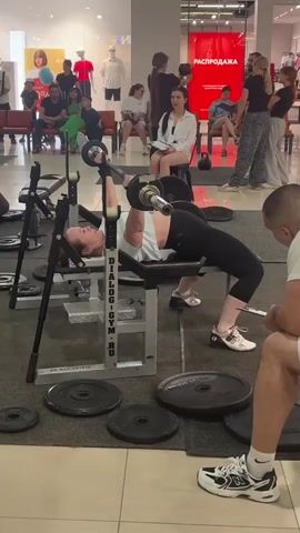 A woman is performing a bench press in a public space, possibly a mall, with spectators watching. She is lying on a bench press machine, with a barbell overhead, and is in the process of lowering the bar. Her form is arched, indicating she is engaged in a weightlifting competition or demonstration. Several people are seated and standing around, observing the exercise. The environment appears to be a retail setting with displays and signage in the background. The woman is wearing athletic attire, black leggings and a white top. The bench press machine has branding that reads "DIALOG-GYM.RU". There are weight plates on the floor surrounding the machine.