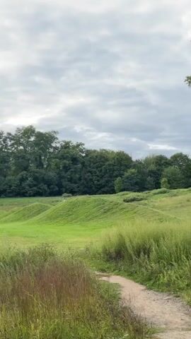 The video shows a gently rolling green landscape under a cloudy sky. In the foreground, tall grass and wildflowers line a dirt path winding to the right. In the midground, several smooth, grassy mounds create a visually striking undulating terrain. On one of the mounds, a small group of people, appearing to be a family, stands together. Behind the mounds and the people, a dense line of dark green trees forms the backdrop of the scene. The overall impression is one of natural beauty and tranquility.