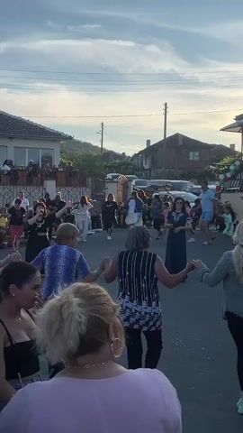 The video shows a group of people dancing in a circle in what appears to be a street. The time of day is late afternoon or early evening, with the sun casting a warm glow and shadows. Several houses and a fence are visible in the background, along with parked cars. The dancers are holding hands and moving in a synchronized manner, suggesting a traditional folk dance. The group is diverse in age and gender, with children also participating. Some onlookers are also present, standing on the side and observing the dance. The atmosphere seems festive and celebratory.