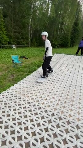 A person wearing a white helmet, white t-shirt, black pants, gloves, and elbow pads rides a snowboard down a ramp. The ramp is made of white circular textured material. The person then rides over a black pipe that is lying on the ground. After going over the pipe, the person slides down a grassy hill on the snowboard. The environment is a grassy hill with trees in the background and a cloudy sky.