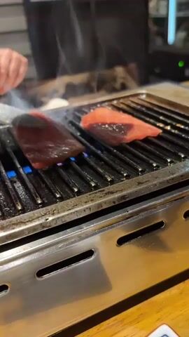 A close-up shot shows a piece of raw red meat being placed on a grill with blue flames beneath the grate. Another piece of meat, already on the grill, is sizzling and smoking. Tongs are used to maneuver the meat on the hot surface. The grill is made of stainless steel with horizontal vents. The background is out of focus, but appears to be an indoor setting with a wooden table.
