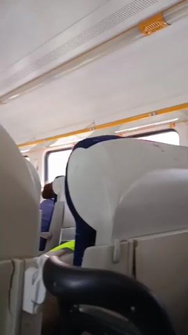 The video shows the interior of a train carriage. The camera is positioned behind a seat, looking towards the front of the carriage. In the foreground, part of a black seat armrest is visible. Further back, a white and blue seatback is prominent. To the left, a person with red hair is seated. A man in a beige jacket, wearing earbuds and carrying a black bag, is walking away from the camera. He is positioned between rows of seats. The carriage has overhead compartments and windows along the side. In the middle ground, a man wearing glasses and a dark shirt is looking down at a phone, with a crossword puzzle book in his lap. To his left, a woman with blonde hair is also looking out the window. Another woman with blonde hair is visible further in the background. The lighting appears to be natural light coming from the windows.