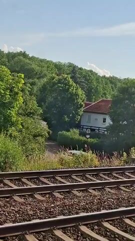 A train track runs along the bottom of the frame. The camera perspective is from inside a moving train. The scene consists of a lush green forest with a small white building nestled among the trees. The building has a red roof and windows. There is a white van parked on a dirt road to the left of the building. The sky is blue with a few white clouds.