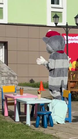 The video shows two costumed characters outside a building on a sunny day. One character is a gray striped cat with a red hat and scarf, and the other is dressed like Pinocchio in an orange shirt and blue pants. They are standing near a small pink and blue table with crayons, a small blue stool, and a speaker. The cat waves his arms as he walks towards a set of stairs. Pinocchio stands next to the table. The building has brown and green sections. A red banner with the word "Альфа Банк" is also visible. The environment includes green bushes, a street light, and an orange cone.