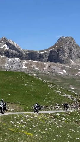 Two motorcycles are riding on a road through a mountainous landscape. The mountains in the background have some snow on their peaks. The road is surrounded by green fields with some rocks and yellow flowers. The sky is clear and blue.