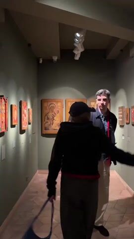 This video shows two individuals in a museum gallery. The gallery walls are painted a dark green, and the floor is made of light-colored wood planks laid in a herringbone pattern. The ceiling is dark and features track lighting. On the left wall, several framed artworks, some appearing to be icons with gold accents, are displayed. In the center of the gallery, two larger framed artworks, also appearing to be icons with religious figures, are prominently displayed. On the right wall, more framed artworks are visible.

The person in the foreground is wearing a black hooded sweatshirt, a black baseball cap, and dark pants. They are holding a blue purse in their left hand and are facing away from the camera, looking towards the artworks on the wall. Their right arm is extended, and they appear to be holding a smartphone, possibly taking a picture or video.

The second individual, a man, is standing to the right of the person in the foreground, facing the artworks. He is wearing a dark jacket with a red collar, light-colored pants, and dark shoes. He is looking towards the artworks and holding a smartphone in his right hand. Both individuals appear to be engaged in observing the art exhibition.