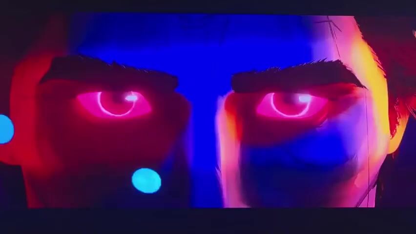 This is a close-up shot of two animated male characters' eyes. They are facing each other, with their faces illuminated by red and blue lighting, giving them a dramatic and intense appearance. The shot then transitions to a view of one character's back as he interacts with a holographic interface. The interface displays various screens with data and images, and the character's hand, clad in a red and blue glove, is seen touching one of the screens. The environment appears to be a futuristic, high-tech setting, suggested by the glowing holographic displays and the complex machinery visible in the background. The character's costume is also red and black, with a distinctive pattern on the back.