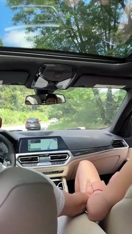 The video shows the interior of a car with two people. A person in the driver's seat has their arm resting on the center console. A person in the passenger seat is wearing sunglasses and has their hand near their mouth. The car is in motion, with trees and a blue sky visible through the sunroof and windshield. The navigation system on the dashboard is active, showing a map.