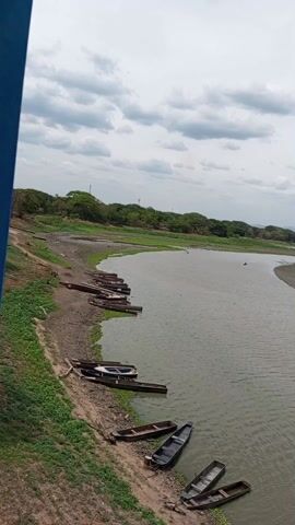 The video shows a wide river under a cloudy sky. The river is bordered by land on both sides, with trees visible in the distance. Several small boats are docked along the shoreline in the lower left of the frame. In the middle of the river, there are several sandbars and what appears to be a few birds. The camera pans from left to right, providing a wide view of the river landscape.
