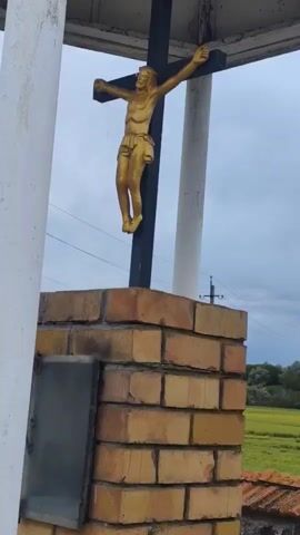 The video shows a brick monument topped with a crucifix. A golden figure of Jesus Christ is affixed to the cross. The monument is surrounded by a field of small, white flowers. The monument is located in an open field, with a cloudy sky in the background. The monument is covered by a white-painted shelter.