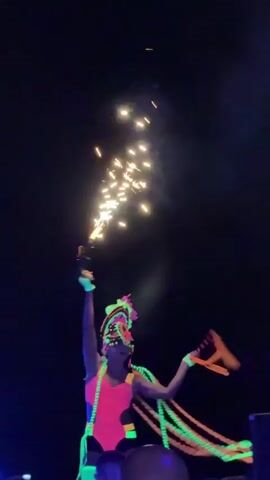 A person in a neon pink outfit with glow sticks is performing on a stage at night. There are fireworks in the air above them. In the background, there is a large, illuminated moon decoration and a crowd of people watching the performance. Some people in the crowd are holding up their phones to record the event. The overall scene appears to be a lively outdoor concert or festival.