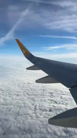 The video shows a view from an airplane window. The wing of the plane, marked with a yellow bird logo, is visible against a backdrop of dense, white clouds below. Above, a blue sky is partially covered with thin, wispy clouds. A contrail from another aircraft crosses the scene diagonally in the upper left.