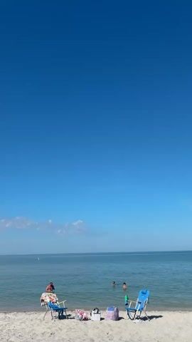 This is a vertical, eye-level shot of a serene beach scene. The bright blue sky takes up the majority of the frame. The horizon line, where the sky meets the calm, blue ocean, is visible in the lower third of the image. A few wispy white clouds are scattered across the sky near the horizon. The foreground consists of a sandy beach. Two blue beach chairs are positioned on the sand, facing the water. In the left chair, a person in a red shirt is sitting. Between the chairs, several bags and a cooler are placed on the sand. Further out in the water, three people are visible swimming. The water is clear and shallow near the shore. The overall impression is one of a peaceful and sunny day at the beach.