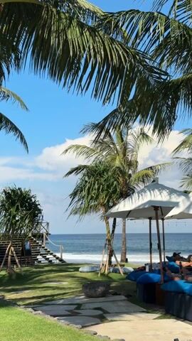 The camera captures a serene scene at a beachside resort, with a framed view through lush palm tree fronds. The turquoise ocean is visible in the background under a clear blue sky with scattered white clouds. In the midground, a manicured grassy area features a wooden staircase leading to the beach, a stepping stone path, and white parasols shading a lounge area with blue cushions. People are lounging in the shaded area, adding a human element to the tranquil setting.