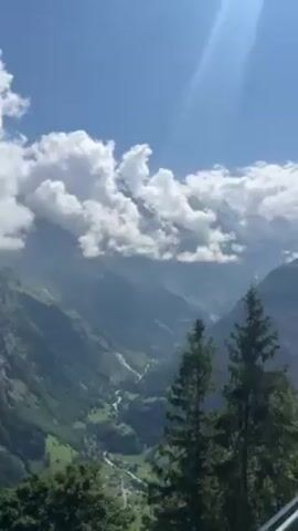 The video shows a picturesque view of a mountain range under a partly cloudy sky.  The mountains are covered in lush greenery, with a winding river cutting through the valley below.  Two tall evergreen trees stand in the foreground, framing the majestic scenery.  The top of one of the mountains is partially obscured by fluffy white clouds, and the sun shines brightly, creating a lens flare effect.  There is a faint sound of voices in the background, suggesting the presence of people, although they are not visible in the frame.